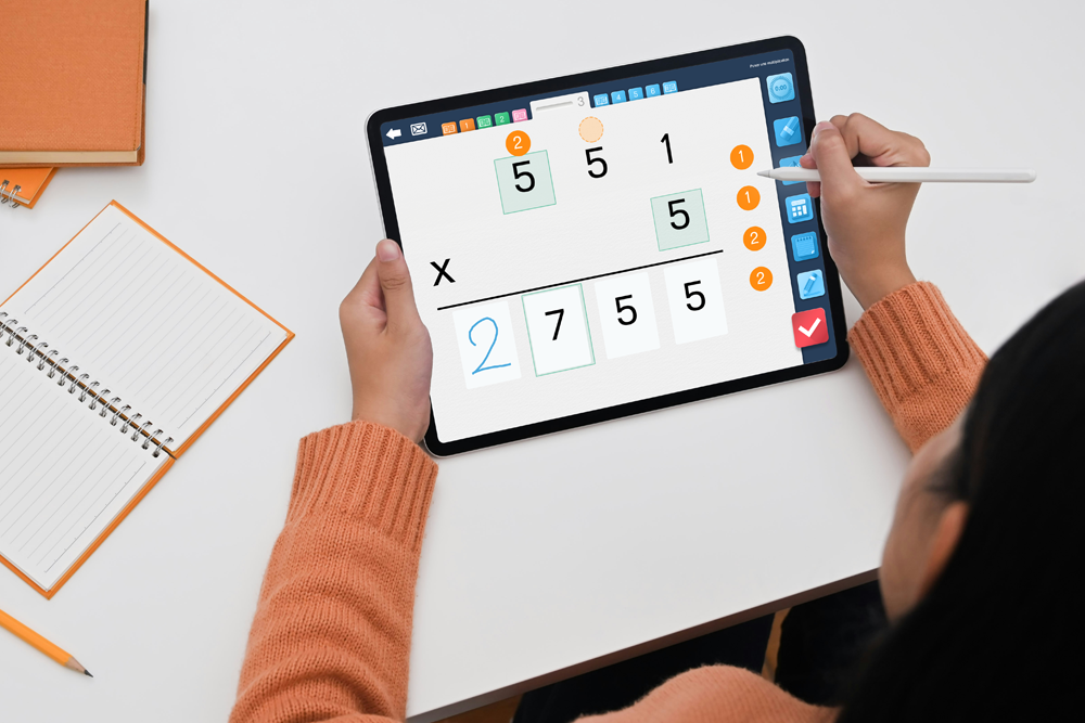 Photo of an iPad displaying a multiplication laid out in the myBlee School app, with a girl completing the exercise. Various notebooks are also on the table.