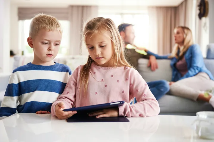 Photo of children, a brother and sister working on an iPad, with their parents talking in the background.